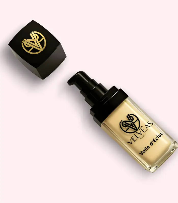 Flacon ouvert Velvéas Voile d'Éclat avec pompe doseuse noire et capuchon avec logo doré posé à côté sur fond rose pâle